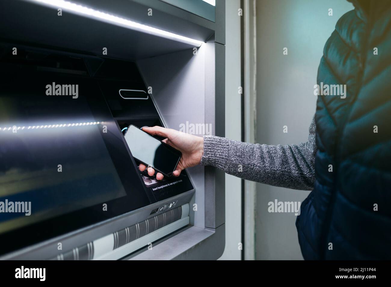 Man`s hand with an smartphone using an ATM street machine to withdraw some cash Stock Photo